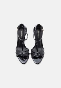 Sandálias de salto alto em couro verniz preto com tiras cruzadas e design em T. Com tira no tornozelo e bico fino. Acabamento brilhante com fivela dourada.