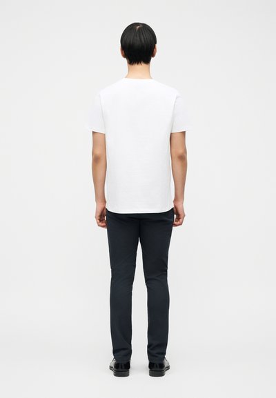 Άτομο που στέκεται με την πλάτη προς τα εμπρός, φορώντας απλό λευκό κοντομάνικο πουκάμισο, σκούρο παντελόνι και μαύρα παπούτσια, μπροστά σε ένα απλό φωτεινό φόντο.