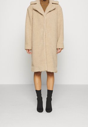 Manteau teddy beige mi-long jusqu'aux genoux porté sur un col roulé rayé, avec des bottines noires, debout devant un fond clair uni.