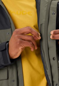 Grüne und schwarze isolierte Jacke mit Reißverschlusstasche, die über einem gelben Hemd getragen wird. Die Jacke verfügt über Druckknopfverschlüsse und strukturierten Stoff.