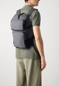 Grauer Rucksack mit mehreren Reißverschlussfächern, rechteckiger Form, gepolsterten Schultergurten und einem verstellbaren Tragegriff. Einfaches Design, keine sichtbaren Muster.