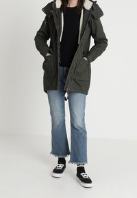 Zelená parka s kapucí podšitou kožešinou, opatřená zipem a zapínáním na knoflíky, dvěma předními kapsami, v kombinaci s krátkými džíny a černými teniskami.