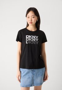 T-shirt nera a maniche corte con logo "DKNY" in argento e motivi di glitter. Abbinata a una gonna di jeans leggera di colore blu. Tessuto di cotone morbido.