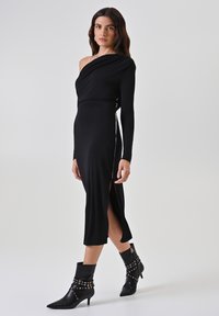 Schwarzes schulterfreies Kleid mit langen Ärmeln und Seitenschlitz. Glatter Stoff mit figurbetonter Silhouette. Getragen mit schwarzen Stiefeletten mit Absatz und Nieten.