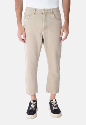 Uomo che indossa pantaloni beige corti e sneaker nere con suole e lacci bianchi, in piedi su uno sfondo chiaro e uniforme.