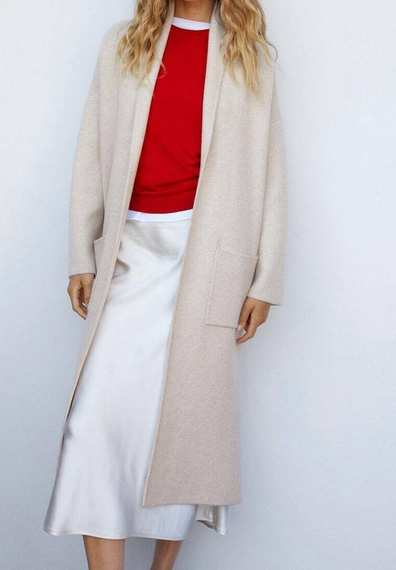 Mujer vestida con un abrigo largo beige sobre un suéter rojo y una falda midi de satén blanca, de pie contra un fondo claro y liso.