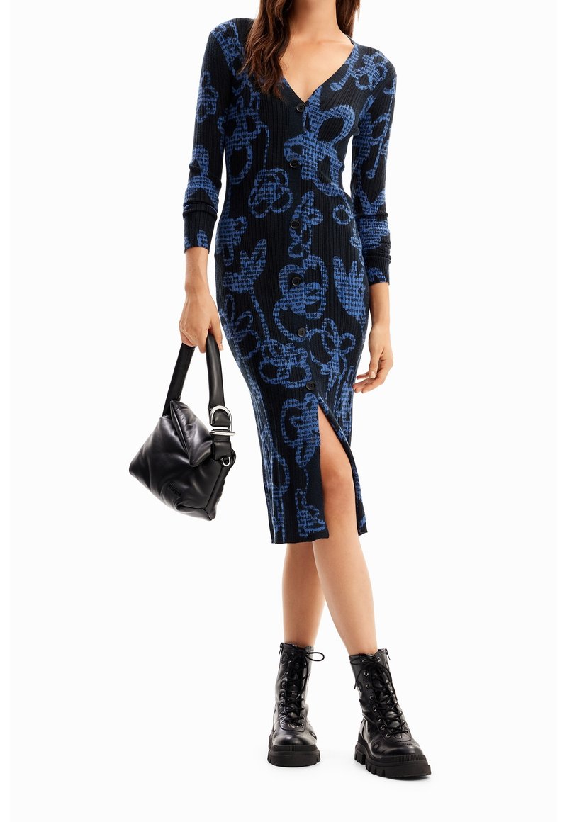Femme portant une robe boutonnée noire et bleue à motifs avec une fente à l'avant, tenant un sac à main noir et portant des bottes noires à lacets.