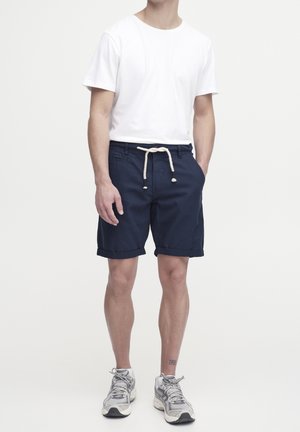 Marineblaue Shorts mit Kordelzugbund, weißem Baumwoll-T-Shirt und grauen Sneakern; die Shorts haben umgeschlagene Bündchen und frontale Taschen.