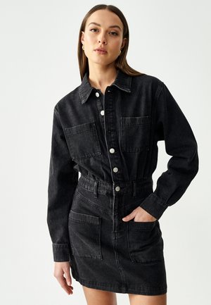 Robe en denim noir avec un col, des boutons-pression et des poches avant. Elle a une taille cintrée et une silhouette de jupe droite.