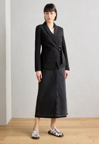 Musta muotoiltu blazer, jossa on kietaisu-design ja hopeinen solki, yhdistettynä pitkään mustaan farkkuhameeseen ja remmisillä varustettuihin hopeisiin sandaaleihin.