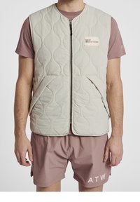 Quilted light beige vest with a front zipper, featuring a label on the chest. Notable textured pattern; side pockets visible. Worn over a pink shirt.