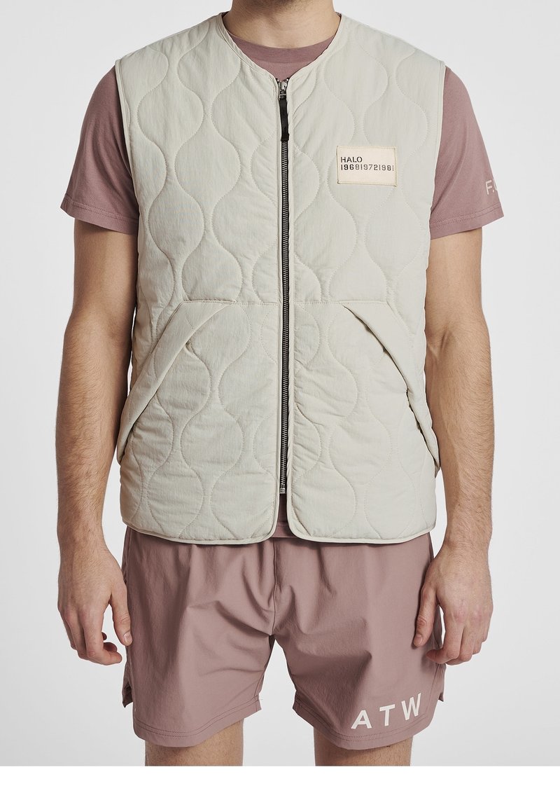 Quilted light beige vest with a front zipper, featuring a label on the chest. Notable textured pattern; side pockets visible. Worn over a pink shirt.