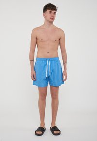 Costumi da bagno blu in materiale leggero e ad asciugatura rapida, con vita elastica e cordino bianco; abbinati a sandali neri infradito.