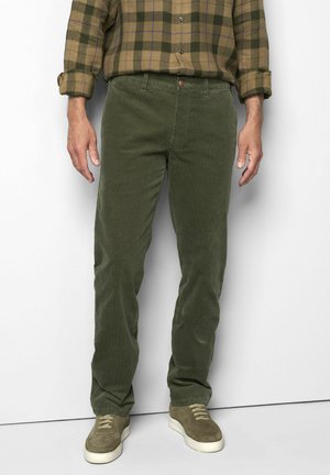 Man draagt olijfgroene ribfluwelen broek, olijfgroen geruit hemd met opgerolde mouwen en olijfgroene sneakers met witte zolen.