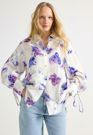 Kvinde med langt blondt hår iført en hvid bluse med lilla blomsterprint og lyseblå jeans, stående med hænderne i lommerne.