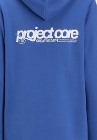 Zils kapucis ar balto drukāto logo, kas lasa "project core" un "CREATIVE DEPT." Teksts ir treknrakstā; materiāls izskatās mīksts un teksturēts.