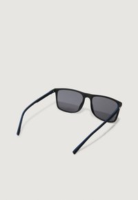 Mustat suorakulmaiset aurinkolasit tummilla sävytyslinsseillä ja kapeilla sinisillä korvissa lepäilevät vaalealla pinnalla.