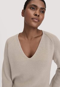 Beige gestrickter Pullover mit tiefem V-Ausschnitt, geripptem Textur und langen Raglanärmeln. Der Stoff wirkt weich und leicht.