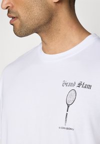 Balta medvilnė marškinėliai su juodu grafiniu atvaizdu, vaizduojančiu teniso raketę, ir tekstu "Grand Slam" senovinio stiliaus. Klasikinis kirpimas.