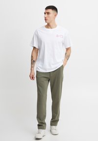 White cotton t-shirt with short sleeves and front text. Paired with olive green trousers and white sneakers, creating a casual look.