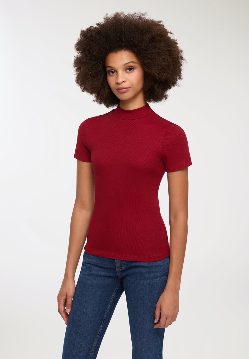 Top rosso a maniche corte con colletto a dolcevita, dal design aderente e texture liscia; abbinato a jeans blu.