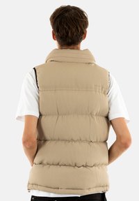 Chaleco acolchado beige con textura acanalada, cuello alto y cierre con cremallera. Lleva puesto sobre una camisa blanca, mostrando un diseño sencillo y funcional.