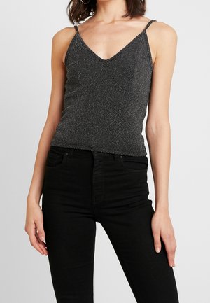 Kobieta ubrana w błyszczącą czarną bluzkę na cienkich ramiączkach i czarne spodnie z wysokim stanem, stojąca na jednolitym jasnym tle.