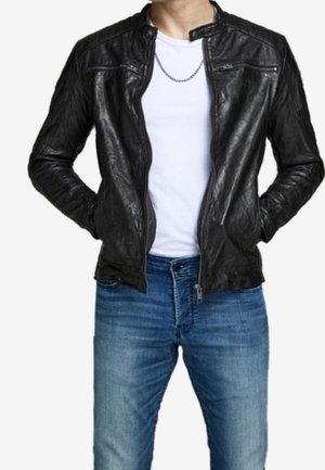 Homme portant une veste en cuir noire par-dessus un t-shirt blanc avec un collier chaîne argenté et un jean bleu, les mains dans les poches de la veste.