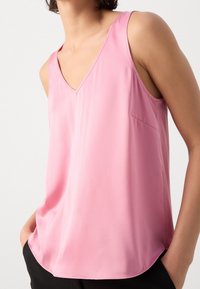 Woman wearing sleeveless pink V-neck blouse with hands in black pants pockets against white background.