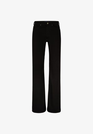 Nainen pukeutuneena mustiin korkeavyötäröisiin bootcut-farkkuihin ja mustiin kärjellisiin nilkkaremmi-korkokenkiin yksiväristä taustaa vasten.