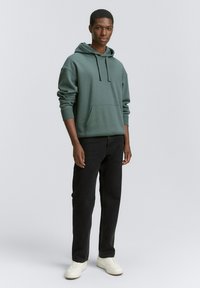 Sudadera verde de mezcla de algodón, con un bolsillo frontal, capucha con cordón, corte relajado, combinada con jeans negros y zapatillas blancas.