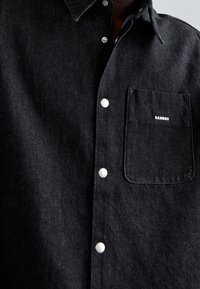 Camicia in denim nero con design a bottoni, bottoni bianchi e una tasca sul petto che presenta il nome del marchio "SANDRO" ricamato in bianco.