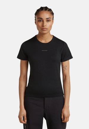 Mujer con el cabello trenzado que lleva una camiseta ajustada negra de Icebreaker y pantalones negros, de pie contra un fondo blanco liso.