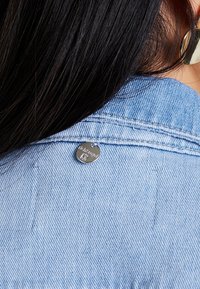 Ljusblå denimjacka med en liten silveretikett vid kragen. Slät textur med synlig sömnad och en klassisk kragsdesign.