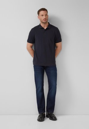 Mann trägt schwarzes Poloshirt, dunkelblaue Jeans und schwarze Schuhe, steht mit verschränkten Händen hinter dem Rücken vor einer grauen Wand.