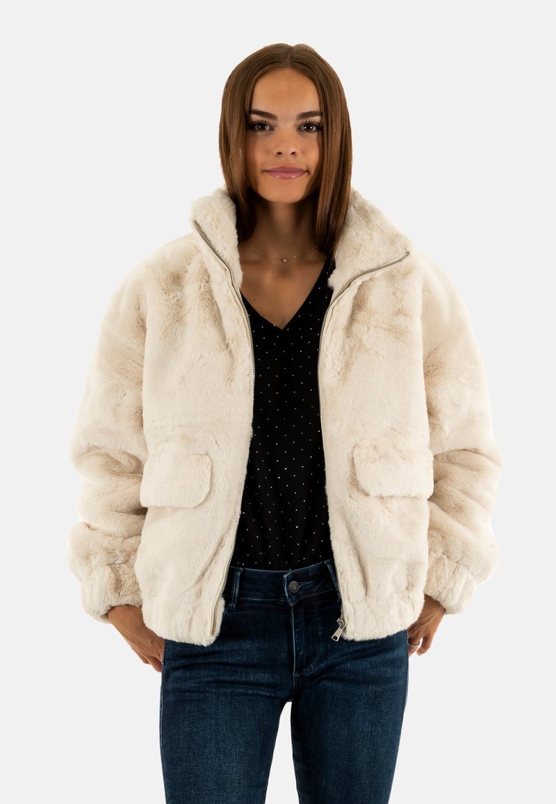 Crème faux bonten jas met ritssluiting aan de voorkant, hoge kraag en twee voorzakken. Draagt over een zwarte blouse met stippen. Zachte textuur, volumineus ontwerp.