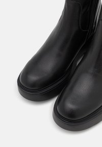 Botas de tobillo de cuero negro con un acabado texturizado, puntera redonda y una suela gruesa negra, que cuentan con un cierre lateral para facilitar su uso.