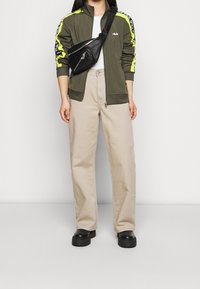 Jasnozielona kurtka na zamek błyskawiczny z neonowymi żółtymi akcentami, zestawiona z beżowymi spodniami o szerokich nogawkach i czarnymi butami na platformie; noszona jest czarna torba na ramię.