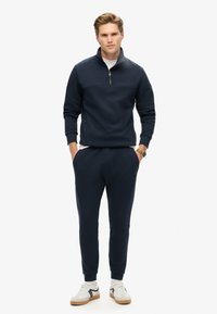 Marineblaues Zip-Hoodie mit hohem Kragen, kombiniert mit passenden Jogginghosen und weißen Sneakern mit schwarzen Akzenten.