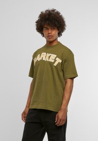 Camiseta de algodón verde oliva con un ajuste relajado, que presenta un audaz texto blanco "MARKET" en el pecho. Mangas cortas y cuello redondo.