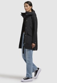 Parka noir imperméable avec capuche, deux poches avant, fermetures à boutons-pression et à zip, porté sur un haut noir et un jean bleu clair.