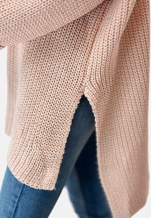 Close-up van een lichtroze gebreide trui met zijopening, gedragen over blauwe denim jeans.