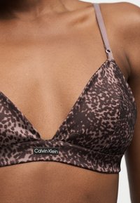 Γυναίκα που φοράει ένα καφέ μοτίβο bralette Calvin Klein με ρυθμιζόμενες τιράντες και ετικέτα με λογότυπο στη ζώνη κάτω από το στήθος.