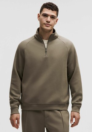 Giovane uomo con capelli corti e ricci che indossa una felpa verde oliva con mezza zip e pantaloni abbinati su uno sfondo chiaro e uniforme.