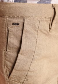 Beige puuvillahousut, joissa on teksturoitu pinta, pleidatut etupuolelta, sivutaskut ja musta "RAW" -logo koristeluna.