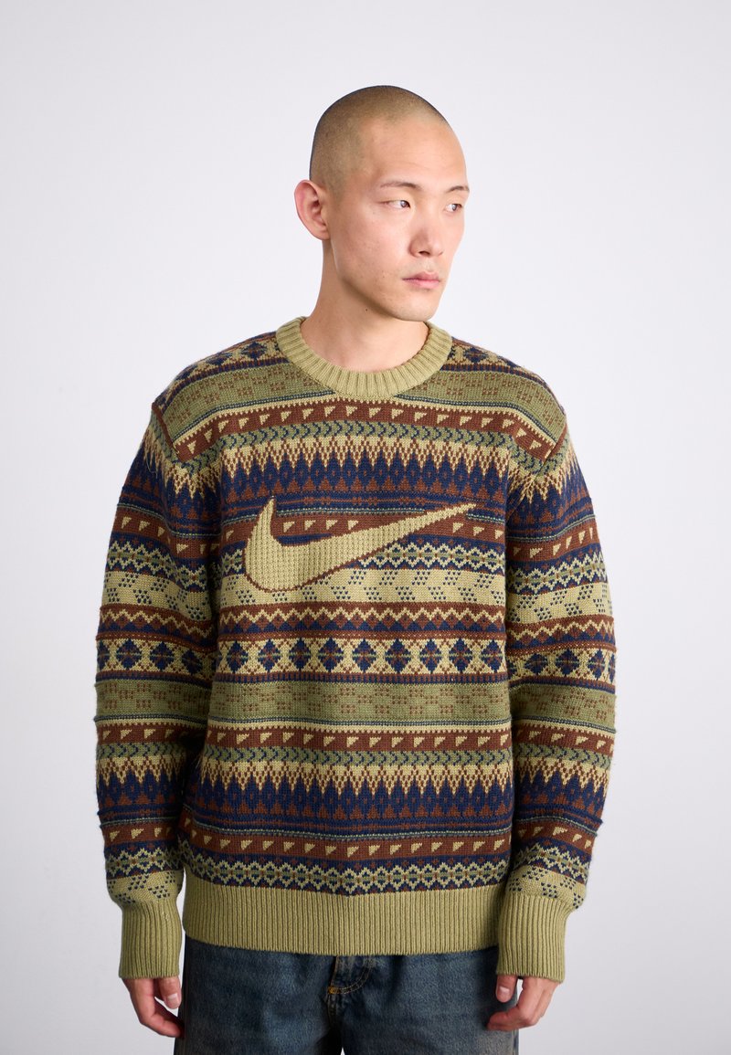Homme avec la tête rasée portant un pull à motifs verts, marron et bleu marine avec un grand logo Nike, se tenant devant un fond blanc uni.