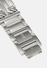 Silver metal bracelet with rectangular links and polished finish. Features engraved "GUESS" logo on clasp and a smooth texture.