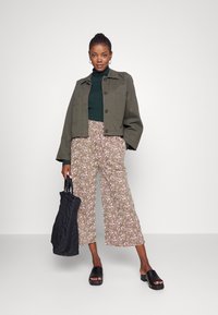 Casaco verde-azeitona cortado sobre um suéter de gola alta verde escura, combinado com calças largas de padrão floral claro e sandálias pretas de salto bloco. Bolsa tote preta.