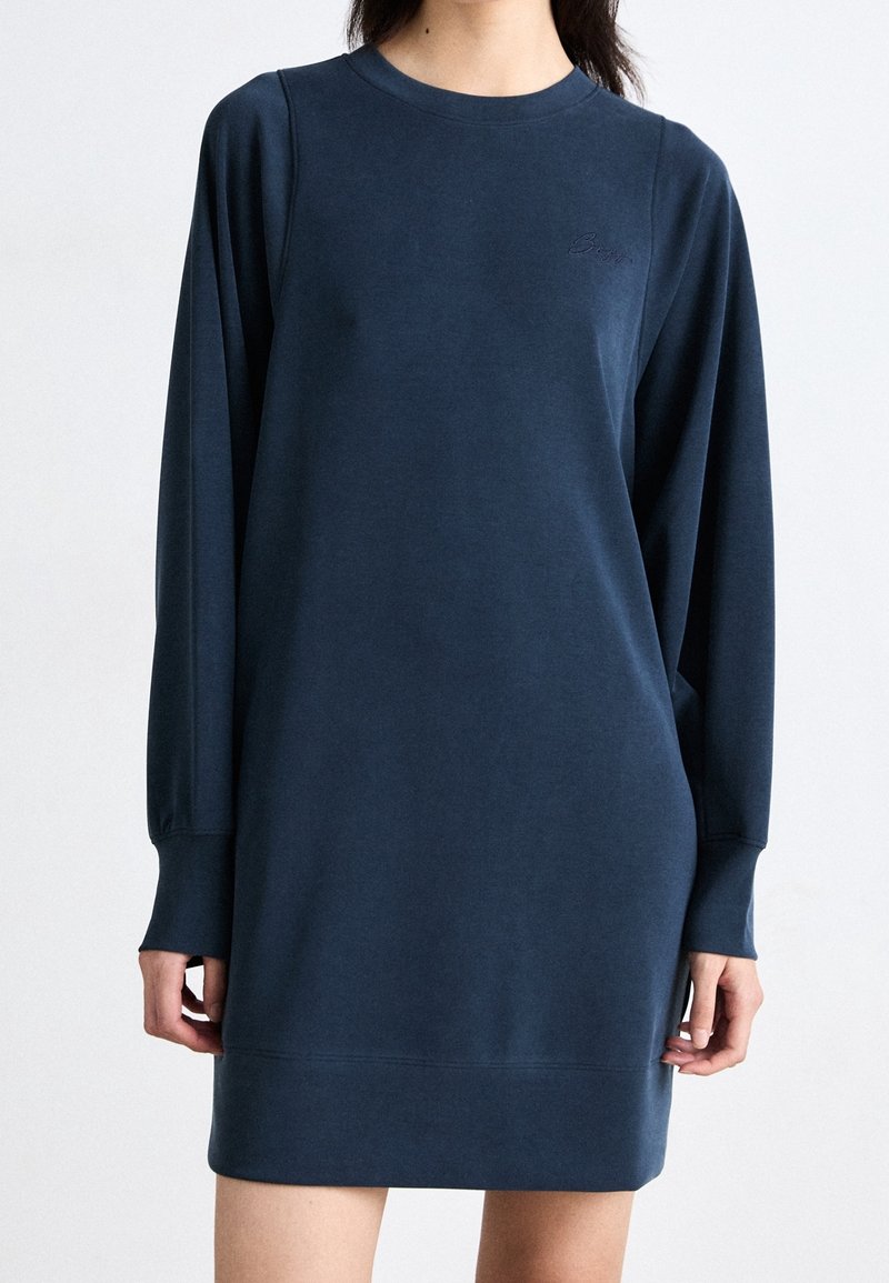 Femme portant une robe sweat bleu marine à manches longues avec un col rond et un logo discret sur la poitrine.