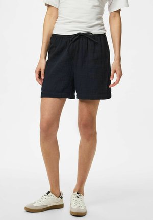 Personne portant un short noir à cordon, un T-shirt blanc et des baskets blanches, debout devant un fond clair uni.